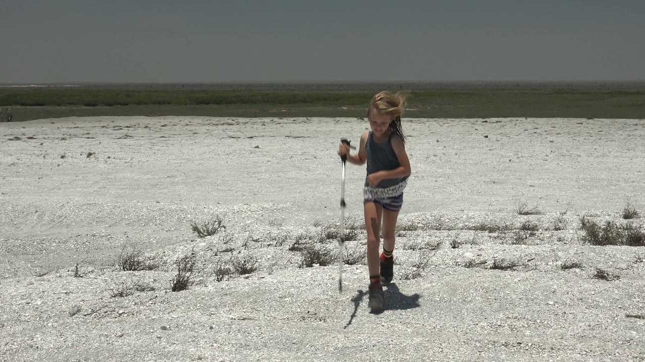 A young girl walks with a stick across a white gravelly plain in San Carlos, Mexico, with sparse scrub and a low horizon behind her.