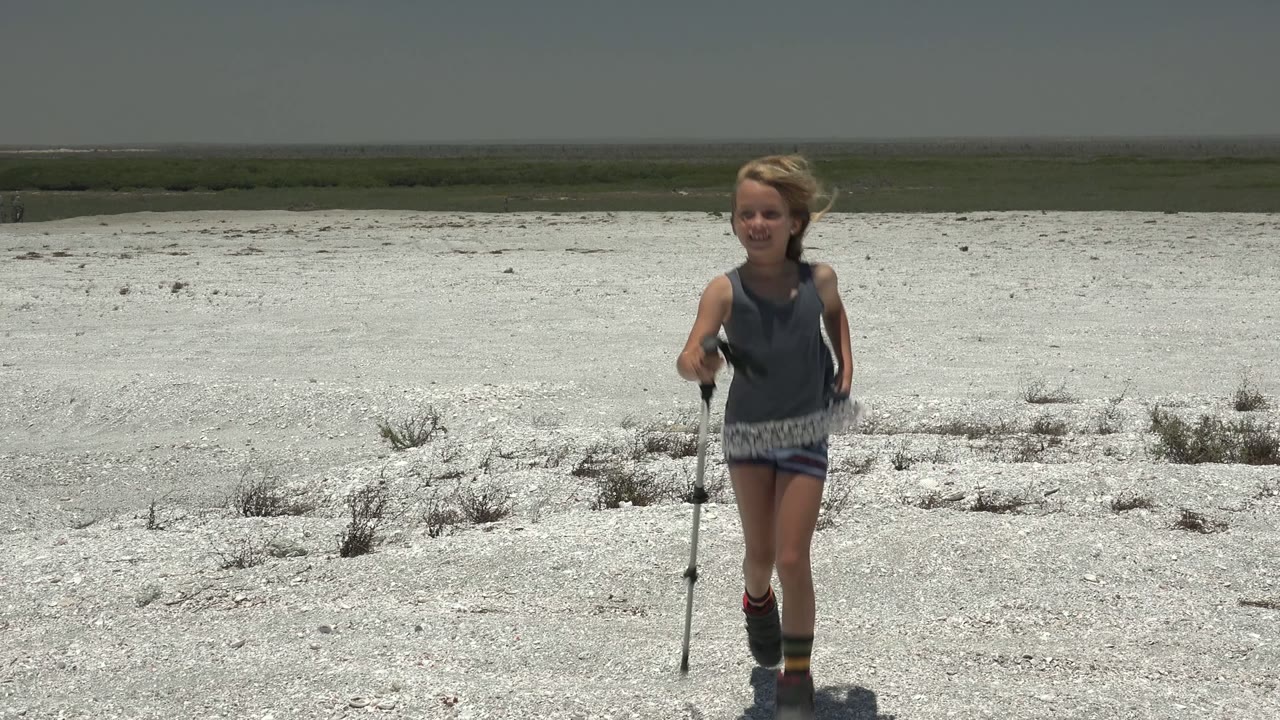 Girl closer to camera while walking across pale desert ground.