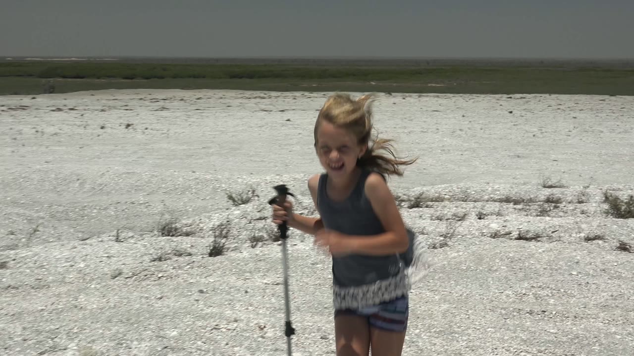 The girl passes close to the camera on the white gravel plain in San Carlos, Mexico, with wind moving her hair and the open landscape behind her.
