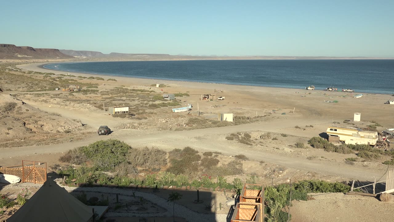 Wide view of San Juanico, Baja California Sur with a sandy road, small vehicles, foreground tent edges, sparse greenery, blue ocean, distant hills, and no visible people.