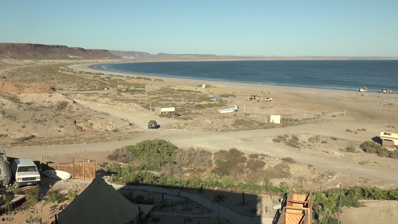 A van appears near the foreground edge in San Juanico as the sandy road crosses the beach area, with camp tents, low shrubs, blue ocean, distant hills, and no visible people.