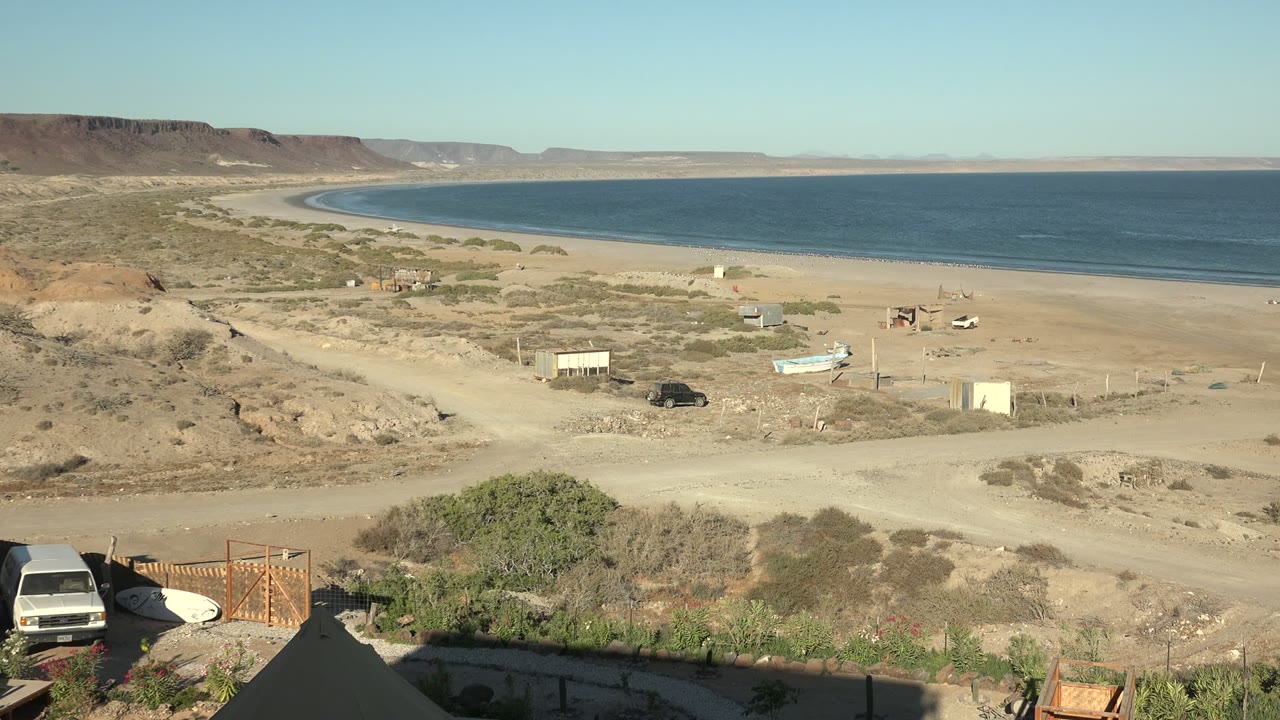 Wide San Juanico coastal desert view with a sandy beach, calm blue ocean, dirt road, white van at the left edge, small boat, scattered structures, distant mesas, and no visible people.