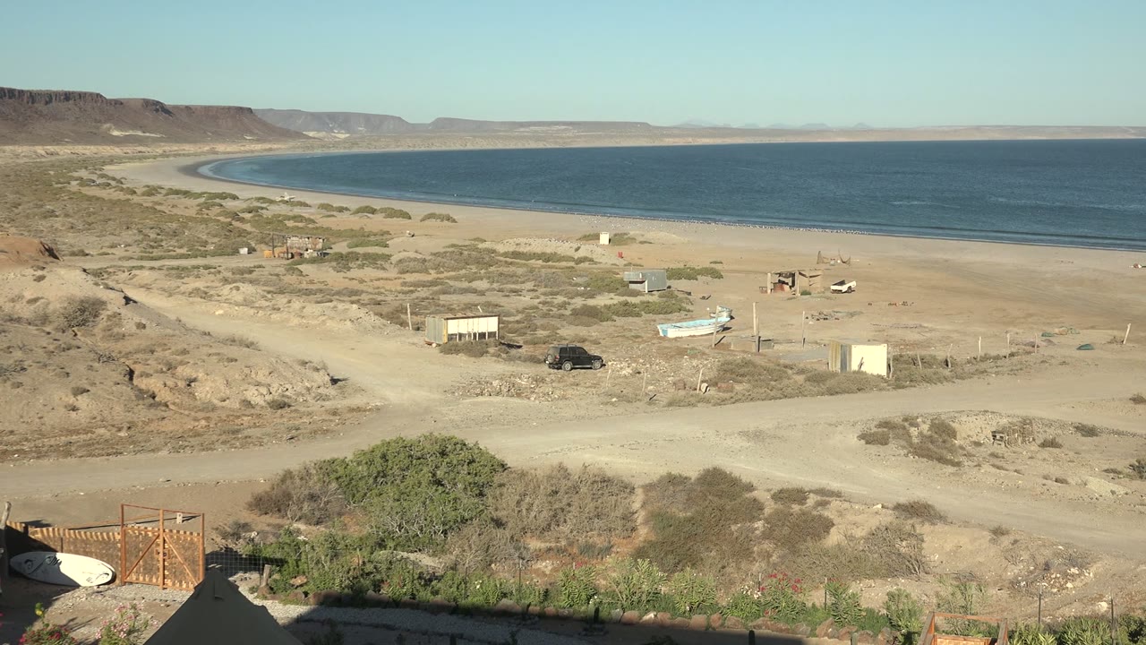 Desert coastline with blue water, sandy beach, dirt road, boat, and small buildings.