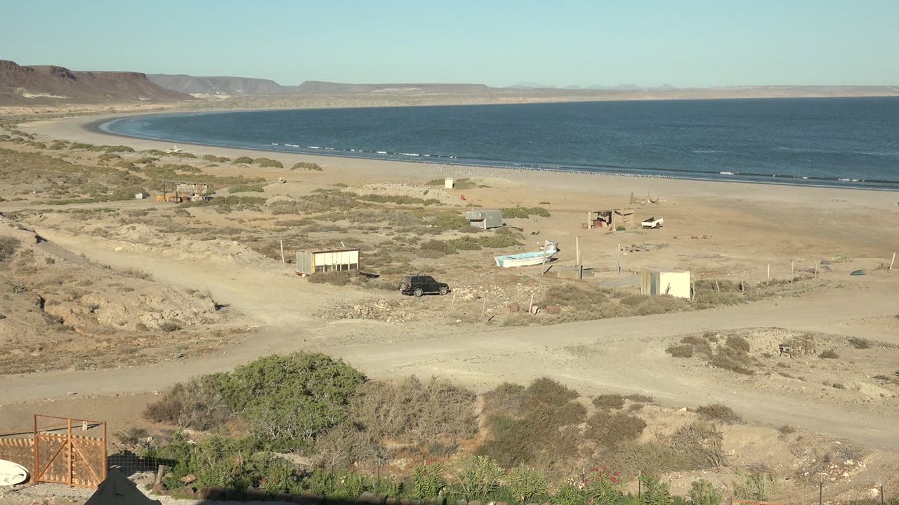 Wide San Juanico shoreline frame with calm water, broad sand, dry scrub, a small boat, a dark vehicle near the dirt road, scattered structures, distant landforms, and no visible people.