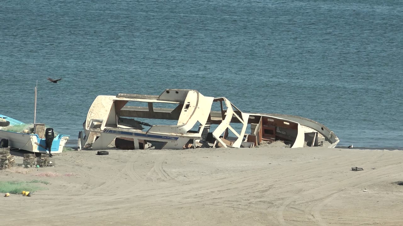 Opening frame in San Juanico, Baja California Sur showing a damaged white boat on sand beside calm blue water, a small blue inflatable boat at left, a bird in the sky, tire tracks, and no visible people.
