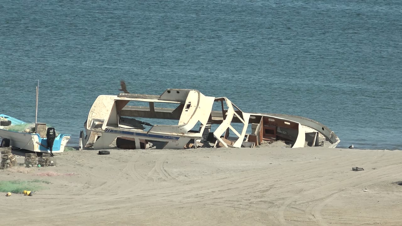 Beached damaged boat and small inflatable boat on sand beside calm water.