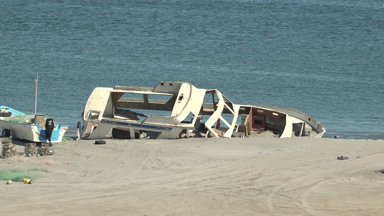Third frame keeps the damaged white boat on the sandy San Juanico shore, with blue water filling the background, beach tracks in the foreground, small objects on the sand, and no visible people.