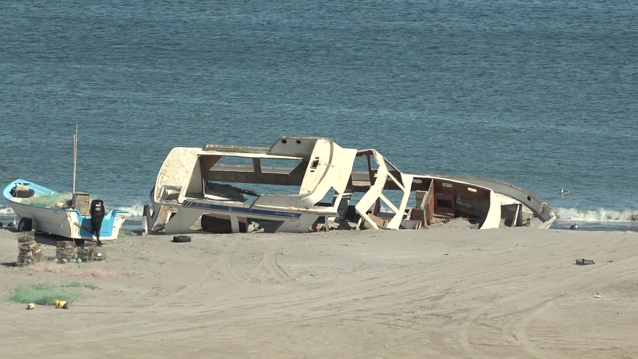 At 0:00 in San Juanico, Baja California Sur, a damaged white and blue boat lies on pale sand beside the ocean with small debris nearby and no visible people.