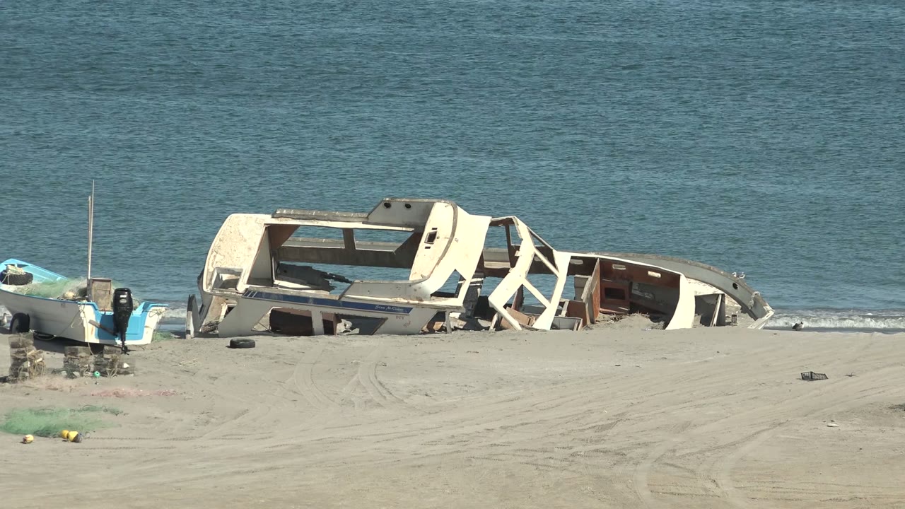 At 0:02, the white wreck sits low along the shoreline in San Juanico, Baja California Sur, with blue water behind it and scattered objects on the sand, no visible people.