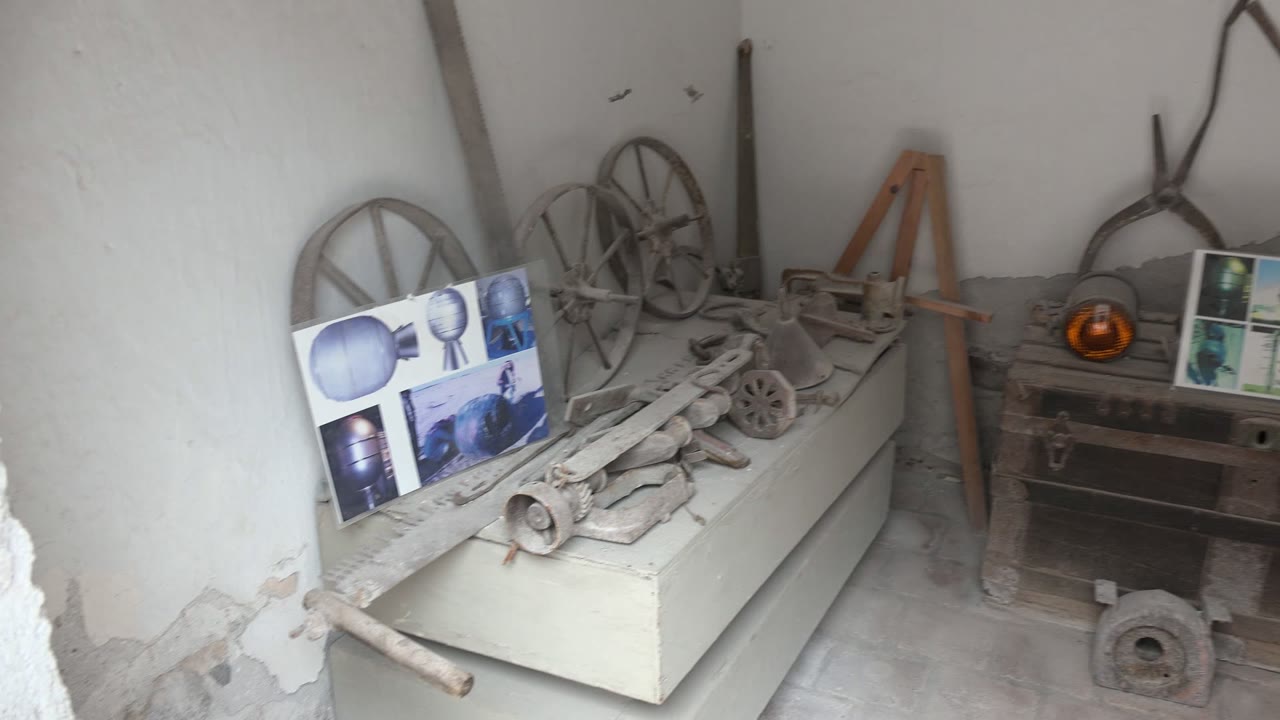 At 0:02 in the Mulege interior, the camera has panned far enough right to include the tool-covered cabinet, wagon wheels, wooden chest, amber object, and a partial display screen with no visible people.