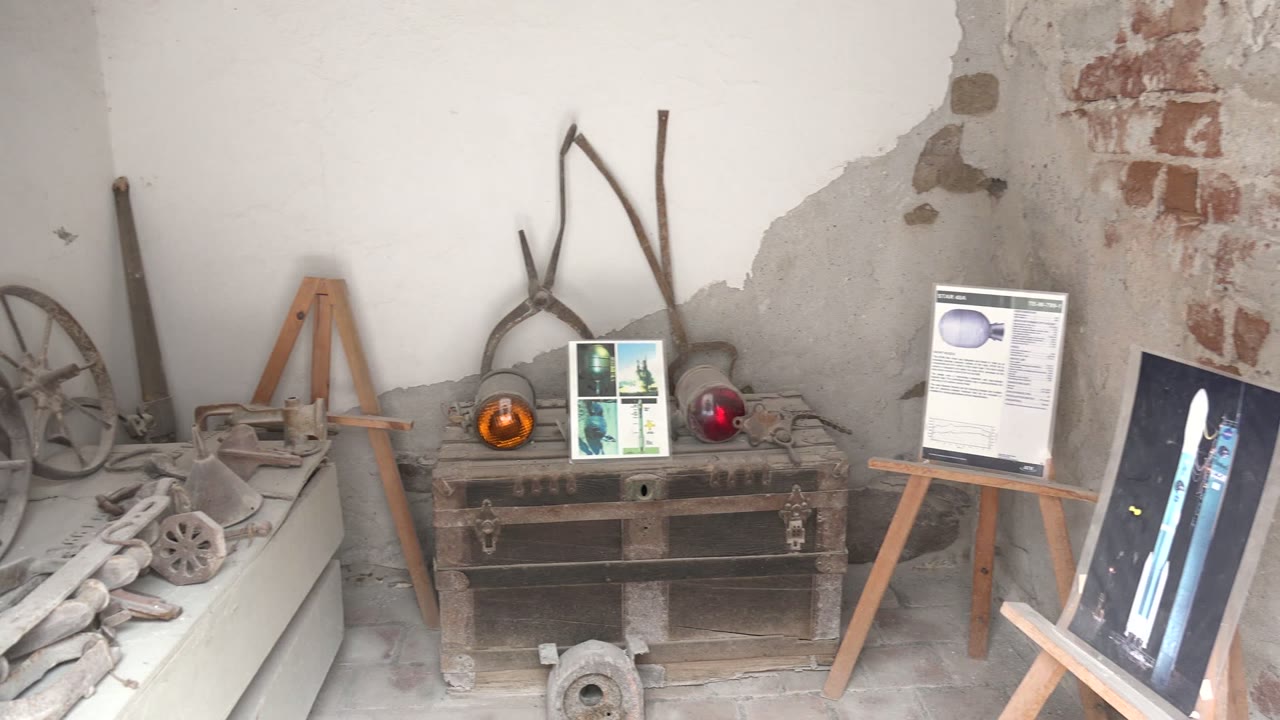 The frame shifts right in the Mulege room, showing less of the tool shelf and more of the easel-mounted boards, wooden chest, red object, and rough wall with exposed brick, with no visible people.