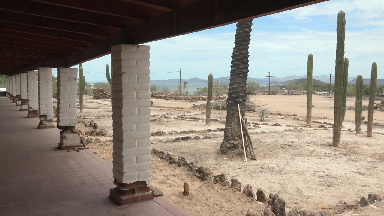 At 0:00, a shaded covered walkway in Mulege, Baja California Sur looks out past white brick columns toward sandy ground, cacti, a palm trunk, distant mountains, and no visible people.