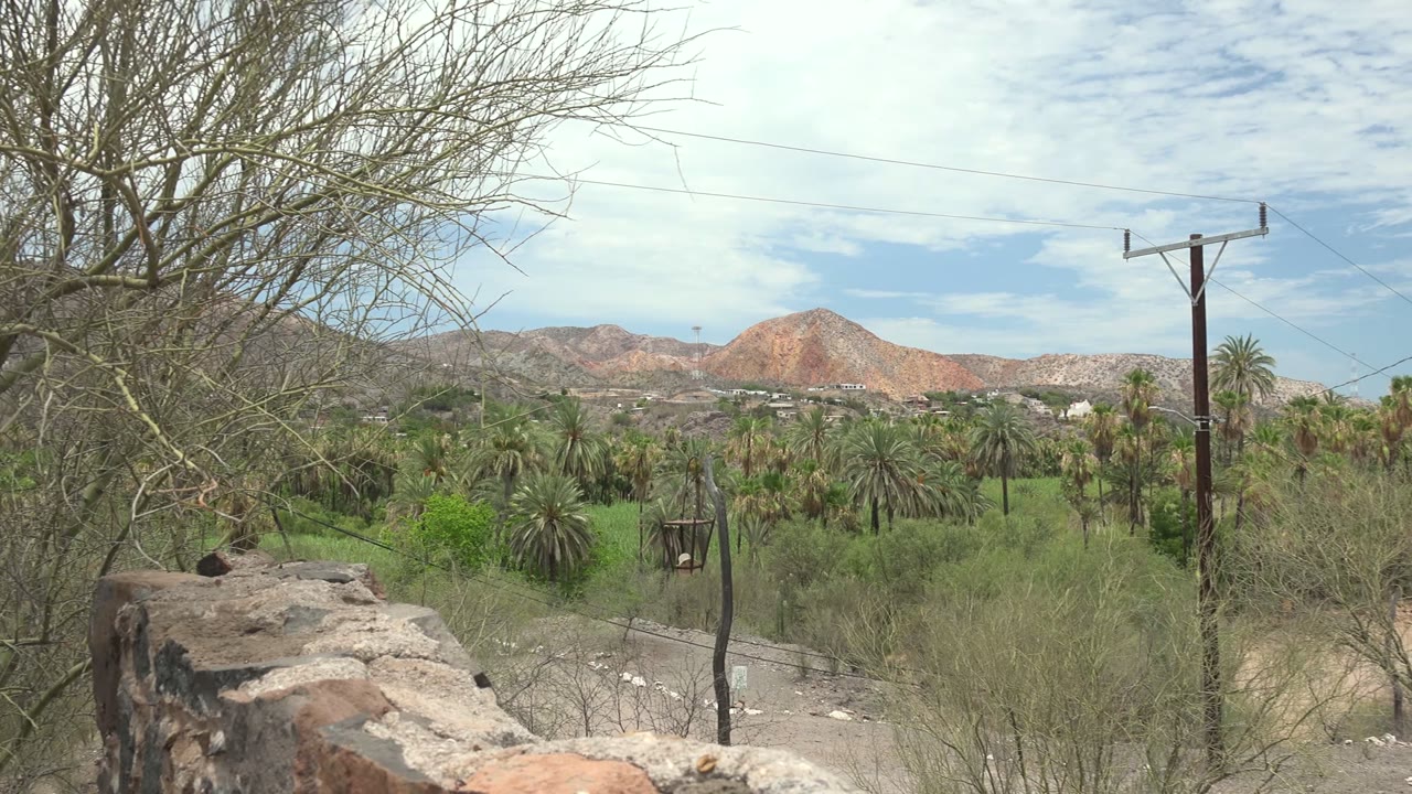 Opening frame of a Mulege, Baja California Sur desert valley with a stone wall in front, dry branches, palms, a power pole, distant reddish mountains, scattered clouds, and no visible people.