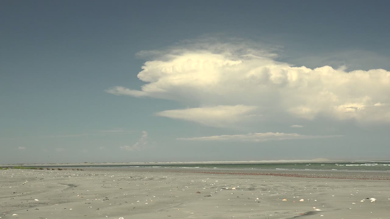 Opening frame of La Bocana, Baja California Sur, with shell-strewn sand in front, a low ocean line, a broad white cloud bank, and no visible people.