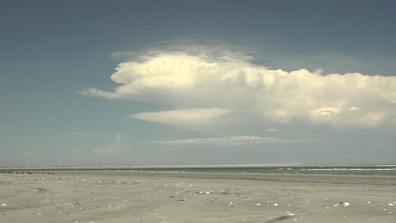 La Bocana beach at 0:02, with the cloud mass stretched over the right side of the horizon, small waves offshore, shell fragments on sand, and no visible people.