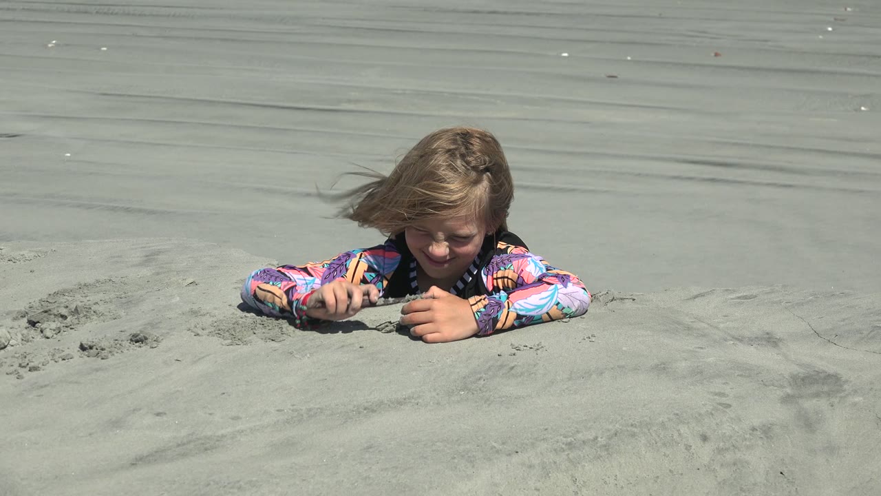 At 0:00 in La Bocana, Baja California Sur, a child lies low in pale wet sand and looks down while digging near a smooth stretch of shallow water.