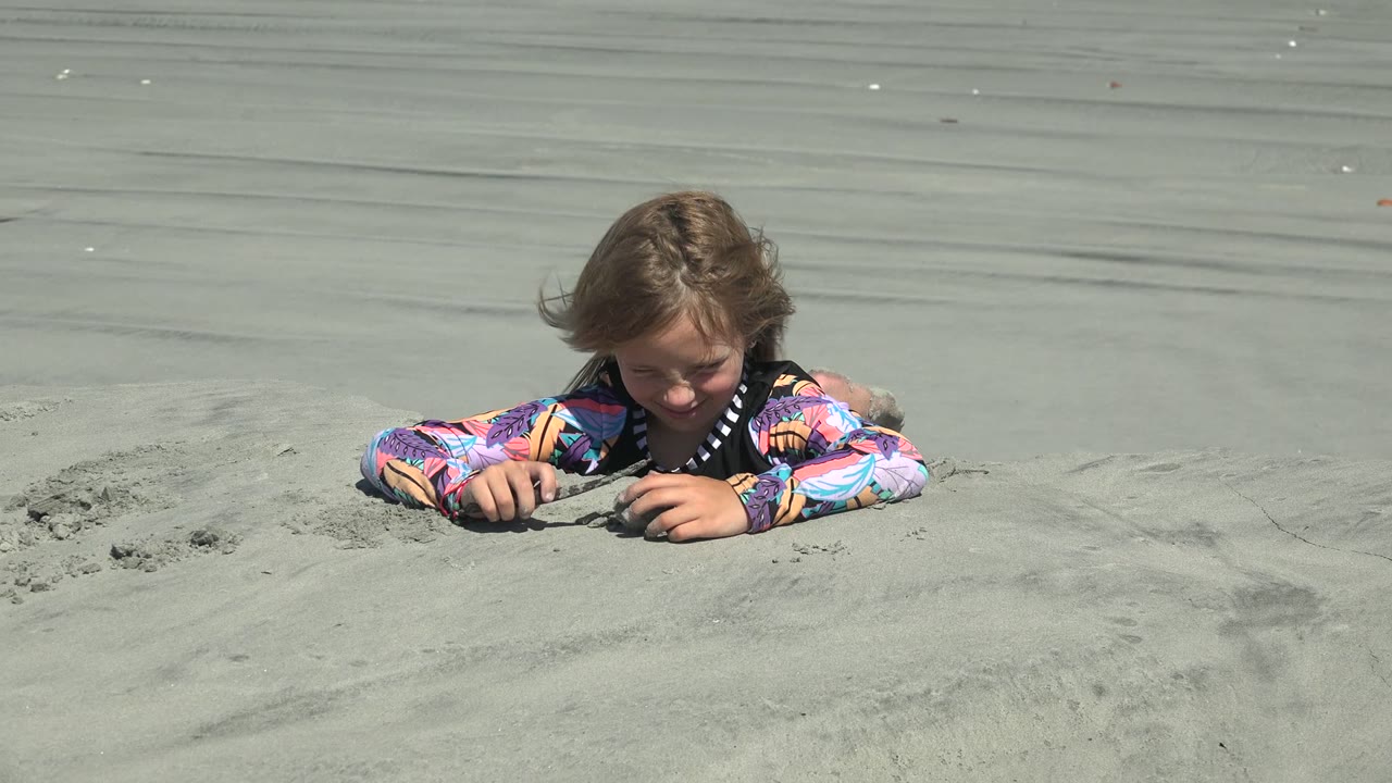 At 0:02 in La Bocana, Baja California Sur, the child looks down toward their hands as both forearms rest on the sand and wind lifts hair across the face.