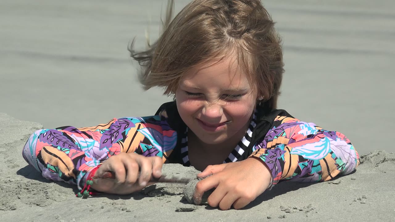 At 0:00 in La Bocana, Baja California Sur, a child in a bright patterned rash guard lies in pale sand with both hands near a small sand shape.