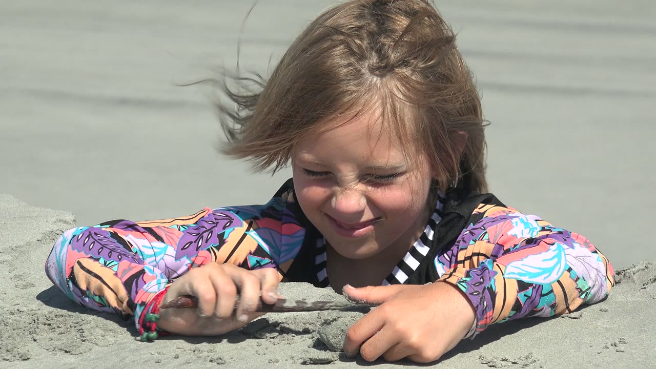 At 0:02 in La Bocana, Baja California Sur, the child leans low over the sand with a hand near a small clump and hair blown sideways across the forehead.
