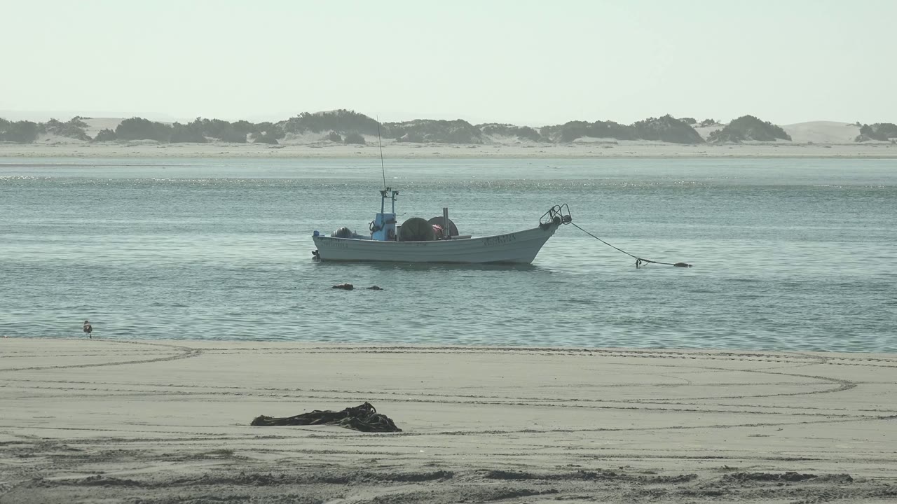 Small boat anchored in calm water off the sandy beach at La Bocana, Baja California Sur, with distant dunes and no visible people on the foreground sand.