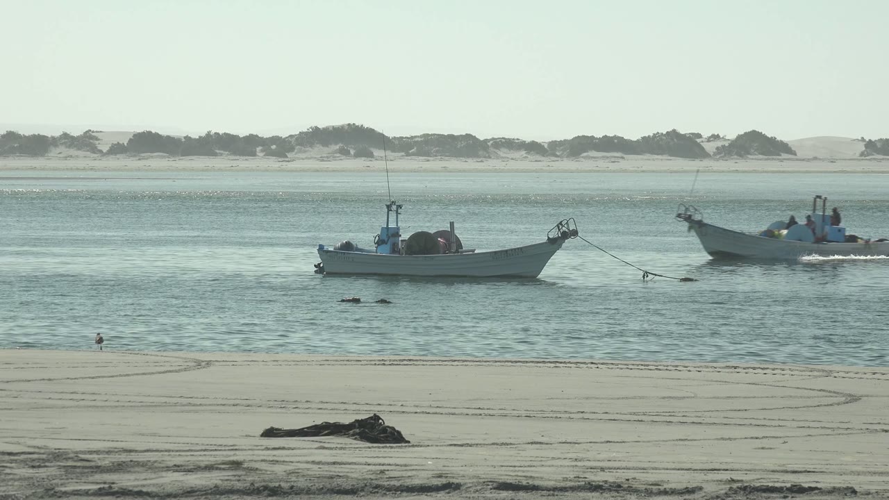 Two small boats share the midground near La Bocana, with the anchored boat left of center, the moving boat to the right, and dunes across the far shore.