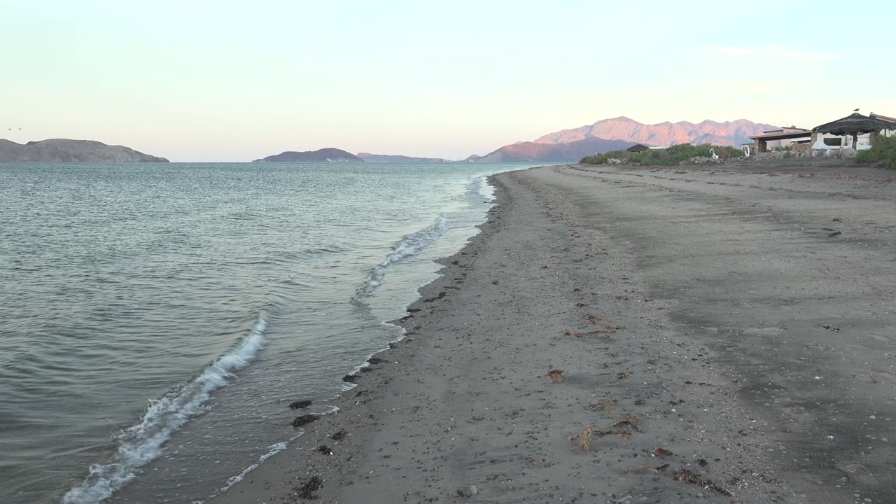 At 0:00 in Bahia De Los Angeles, gentle surf runs along the left edge of a sandy beach, with low islands and pink-lit mountains in the distance and no visible people.