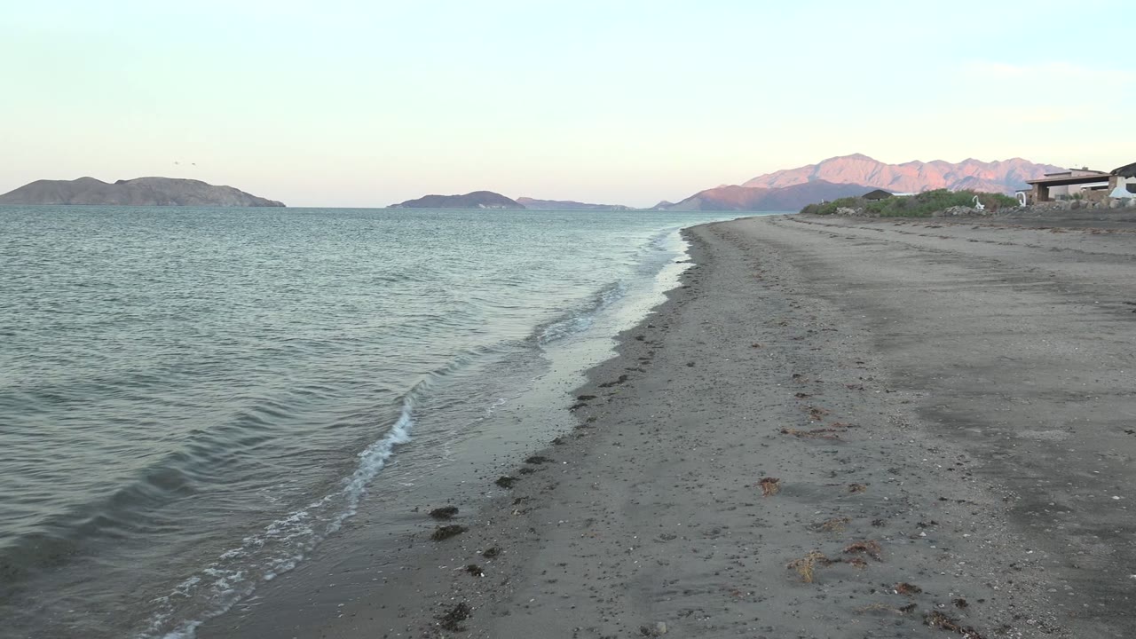 At 0:02 in Bahia De Los Angeles, rippled teal water presses against the sand, with the beach curving toward pink-lit mountains and no visible people.