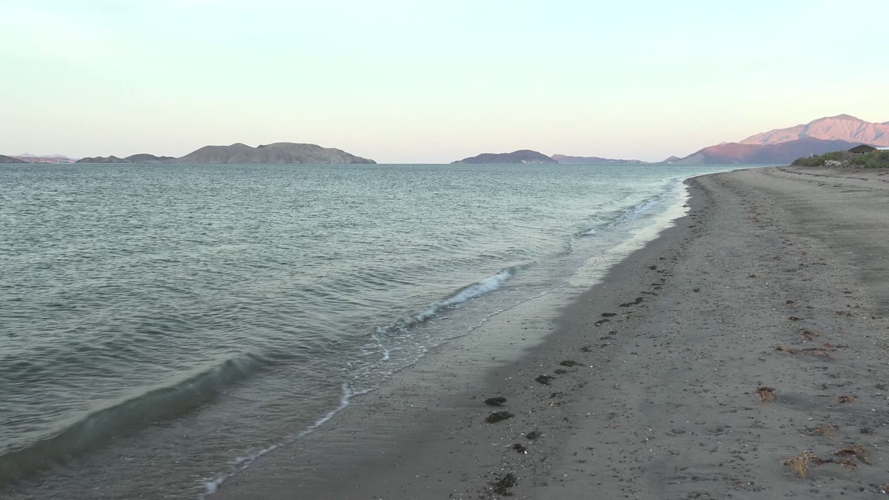 Opening frame of Bahia De Los Angeles shoreline with small waves along wet sand, footprints leading away, distant mountains on the horizon, and no visible people.