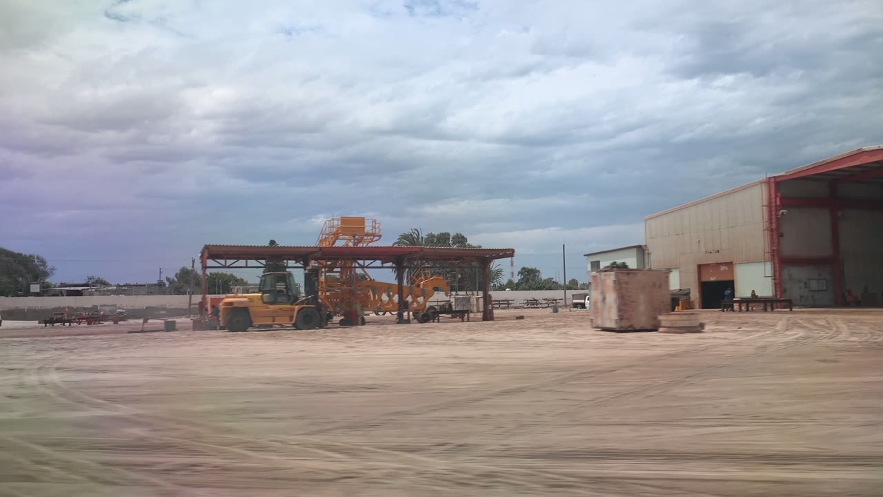At 0:00 in Guerrero Negro, Baja California Sur, a wide dusty industrial yard shows orange heavy equipment near a steel canopy and a warehouse, with no visible people.