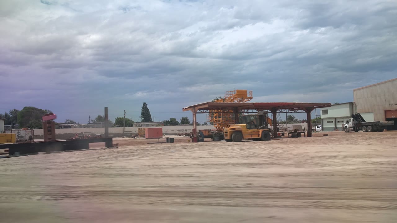 At 0:02 in Guerrero Negro, Baja California Sur, the pan reveals a warehouse side, a parked truck, low yard structures, and heavy equipment under cloud cover, with no visible people.