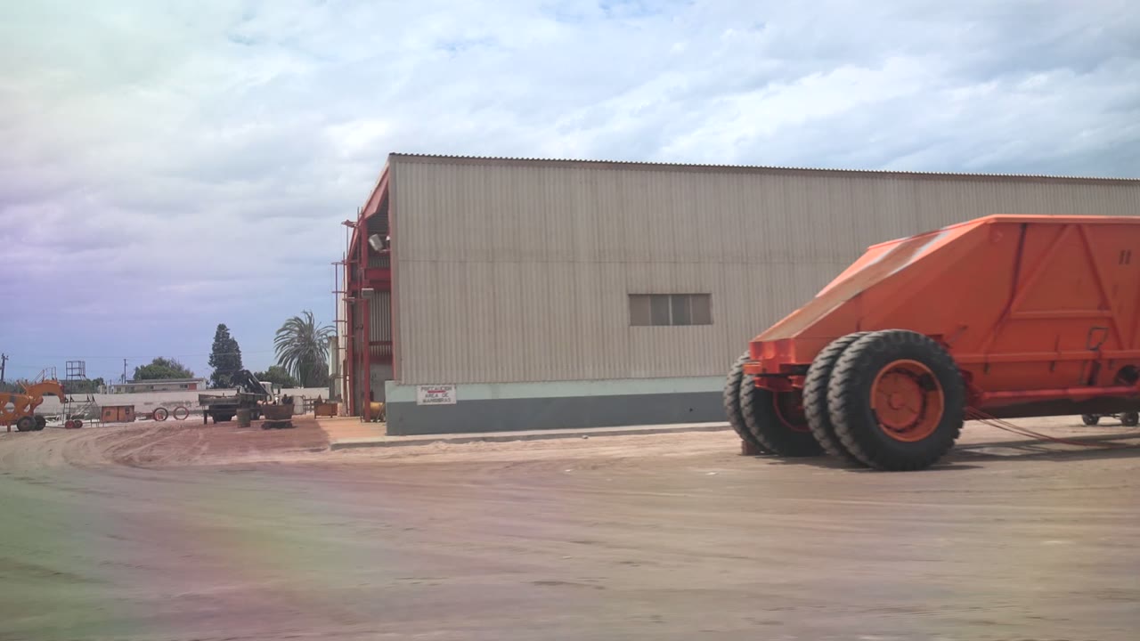 Opening view of a Guerrero Negro industrial yard with a beige warehouse, red framing, orange heavy machinery at right, open concrete and dirt ground, cloudy sky, and no visible people.