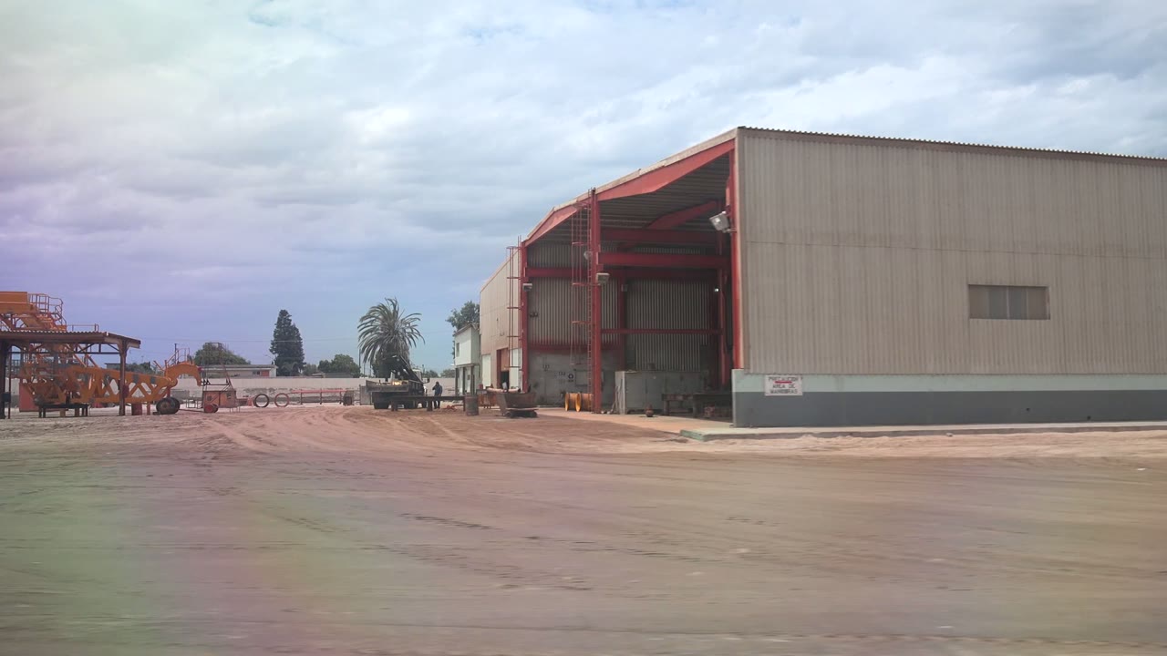 A wider view of the Guerrero Negro industrial yard centers the warehouse's red-framed open bay, with machinery scattered across the lot, a palm tree in the distance, cloudy sky, and no visible people.