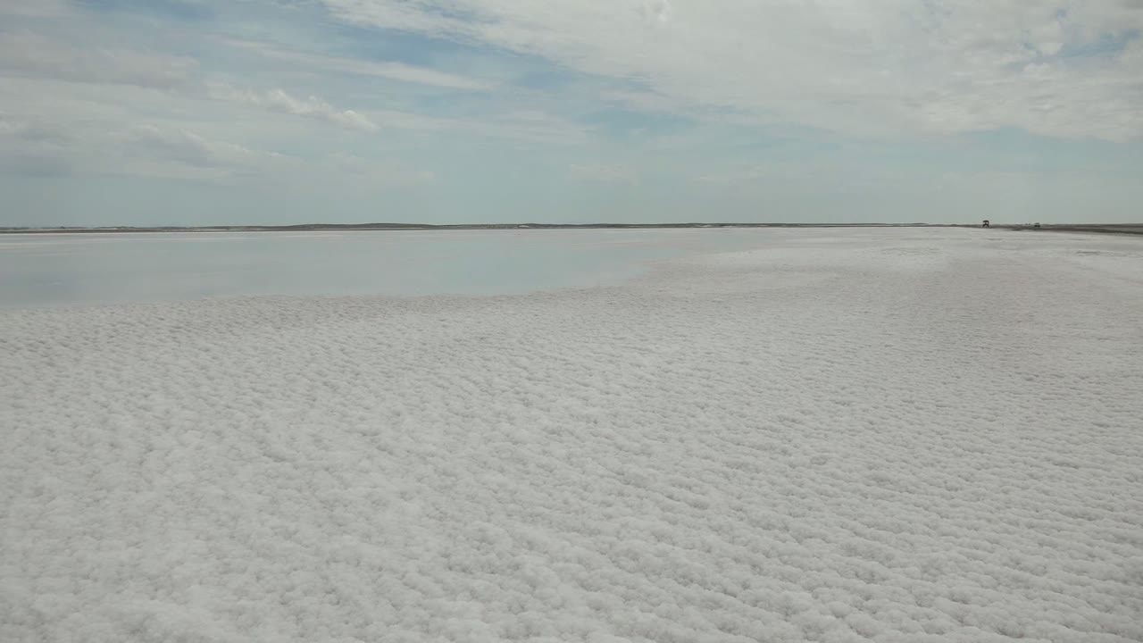 Opening view of Guerrero Negro salt flats with rough white foreground texture, a narrow calm water band, distant low shoreline, pale clouds, and no visible people.