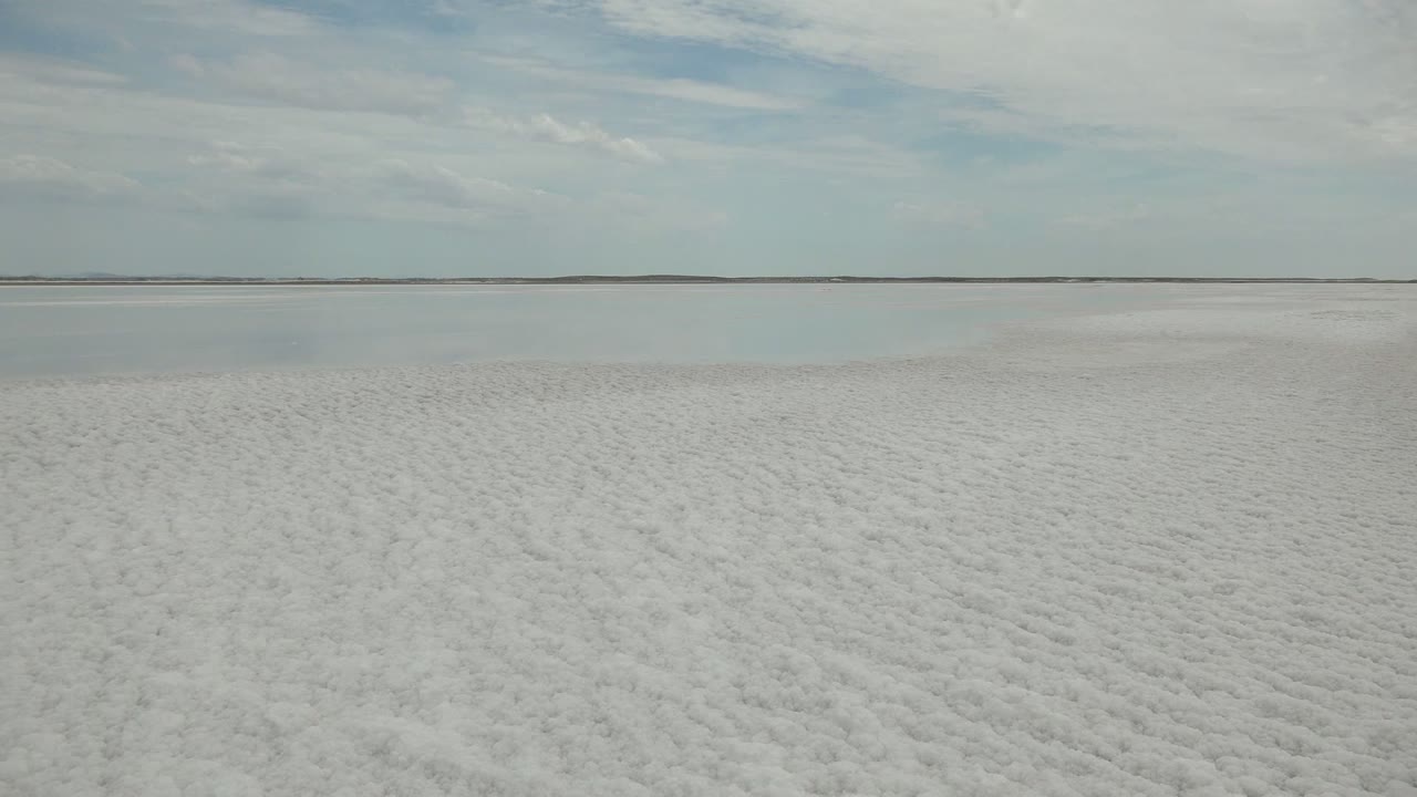 A pale white salt expanse in Guerrero Negro stretches to a smooth water band and distant low land under light cloud cover, with no visible people.
