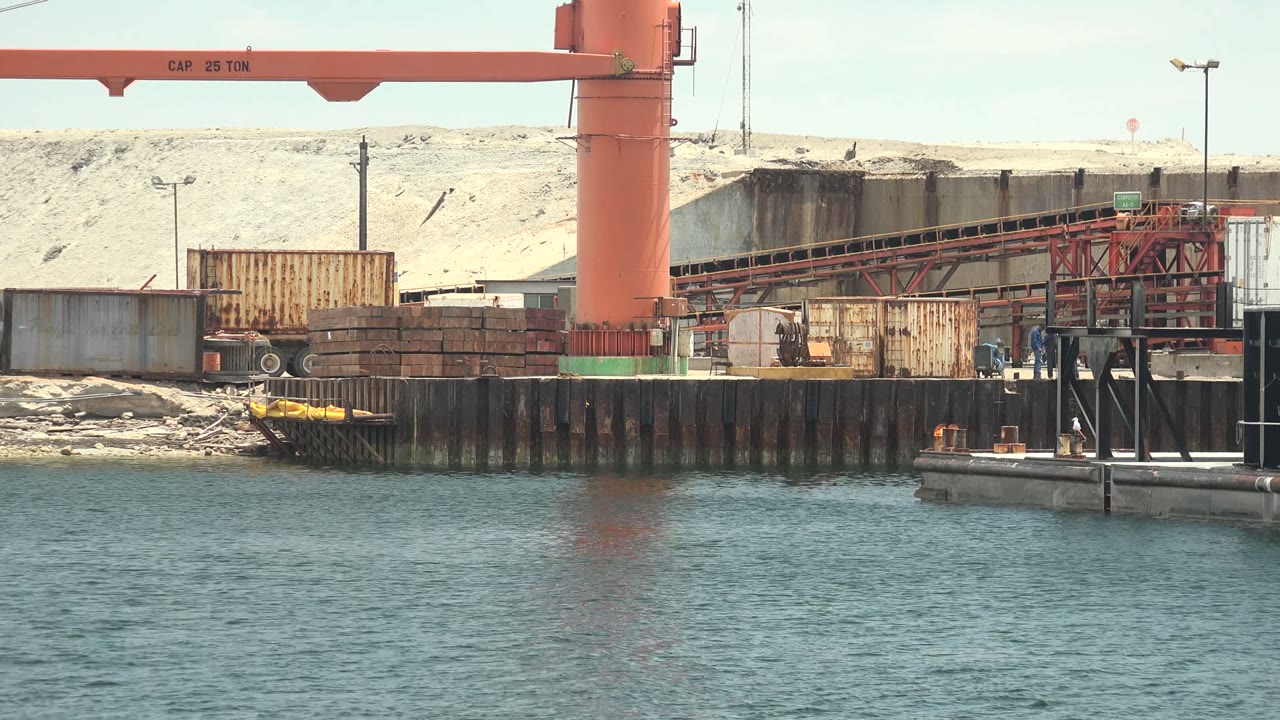 Red industrial crane on a Guerrero Negro dock above calm blue-green water, with containers, a conveyor, pale material piles, and no visible people.