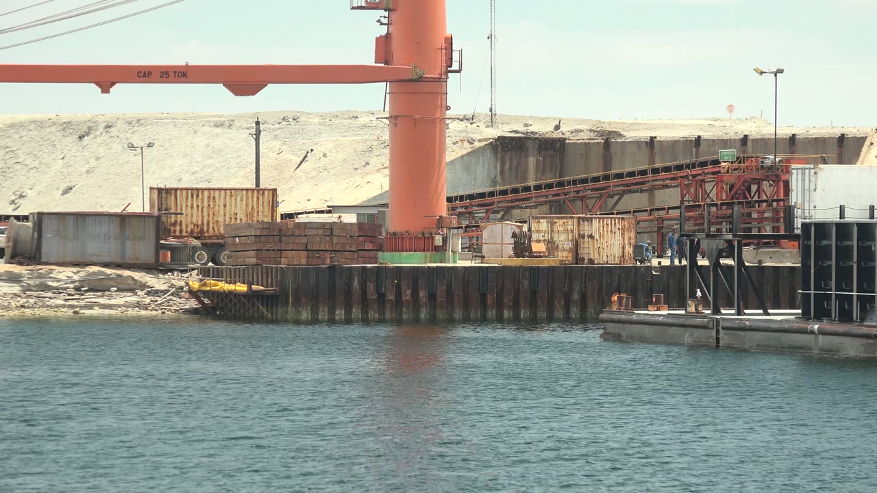 A Guerrero Negro dock scene shows the red crane beside containers and conveyor equipment, with rippled water in the foreground and no visible people.