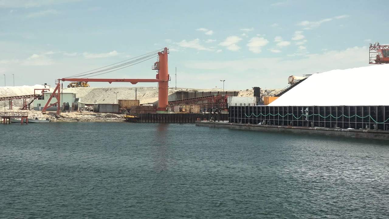 Wide view across calm water toward an industrial port in Guerrero Negro, Baja California Sur, with red cranes, white storage piles, dark dock supports, and no visible people.