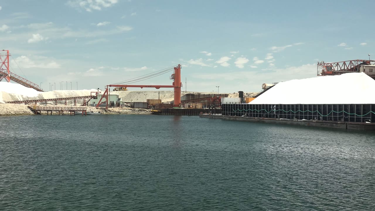 A large white storage mound dominates the right side of the Guerrero Negro port view, with dark dock supports below, red port machinery behind it, calm water in front, and no visible people.