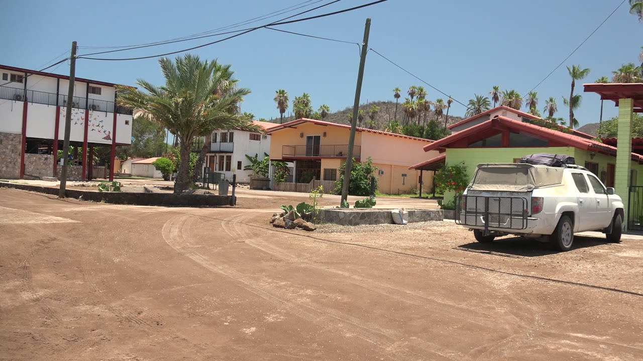 Opening frame of a Mulege, Baja California Sur dirt road with residential buildings, palm trees, overhead wires, and a white pickup partly at the right edge, with no visible people.