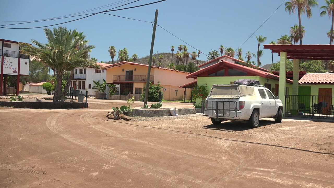 Rightward pan frame in Mulege showing the white pickup near a gated green building, red rooflines, dusty road foreground, palms behind the homes, and no visible people.