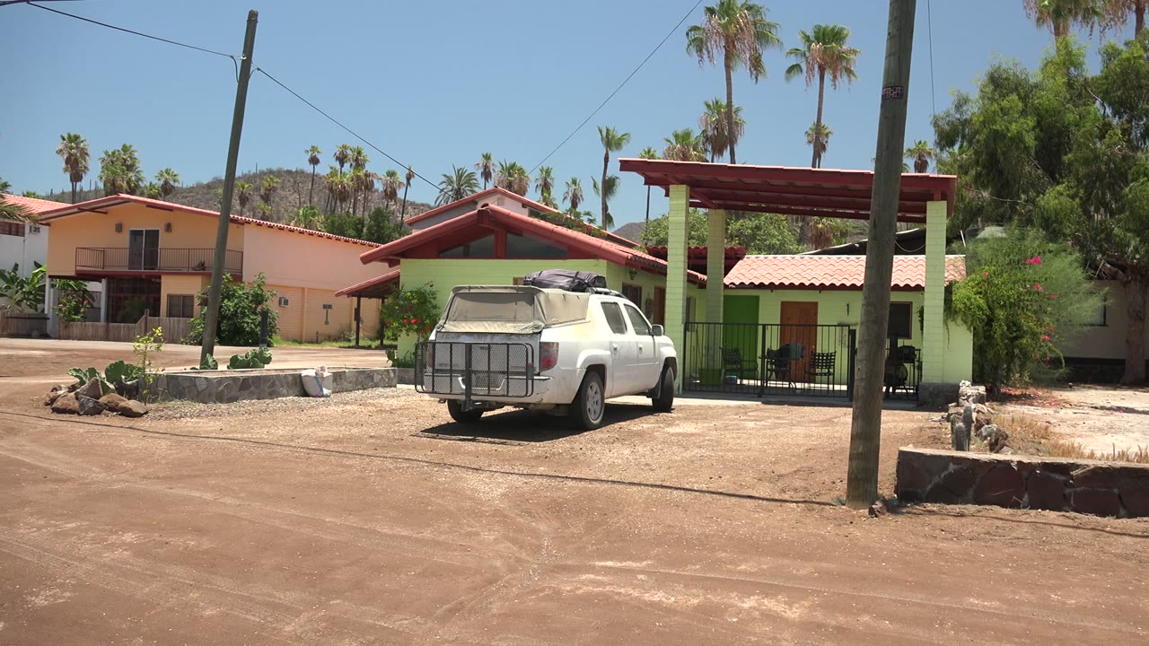 Opening frame in Mulege, Baja California Sur, with a white pickup parked near peach and green houses, palm trees, utility poles, dry dirt ground, and no visible people.
