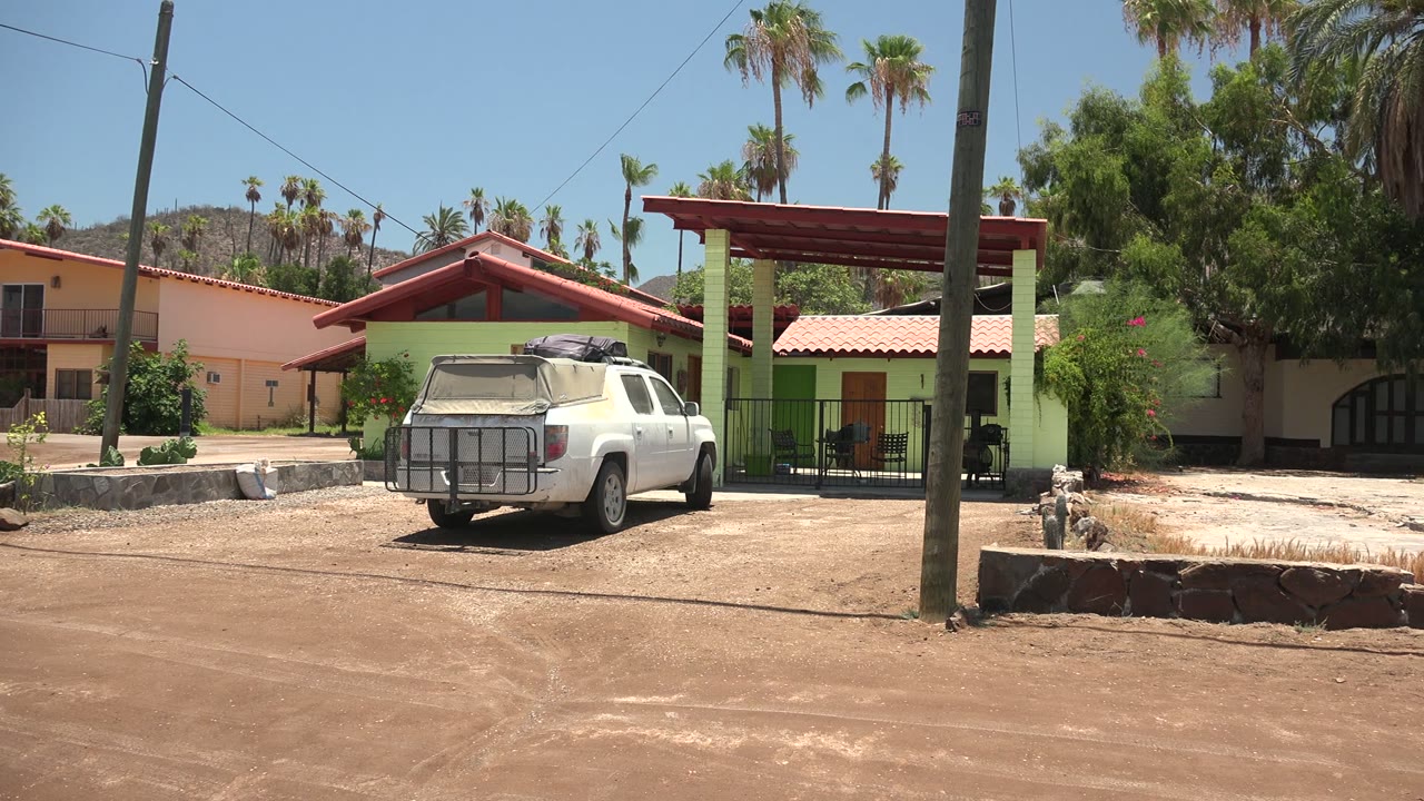 Mid pan frame in Mulege with a foreground utility pole, a white pickup parked beside the green house, peach building edges, palms, dry open ground, and no visible people.