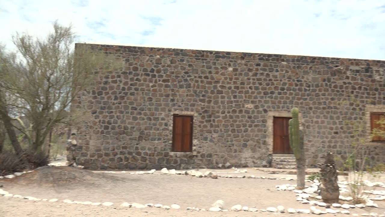 Wide view of a Mulege, Baja California Sur stone courtyard wall with wooden doors, cactus plantings, pale ground, blue sky, and no visible people.