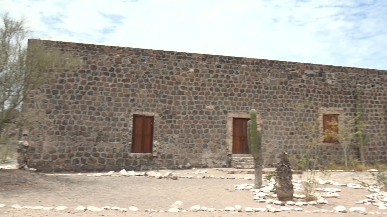 Long stone building wall with wooden doors, cactus, rocks, and dry courtyard plants.