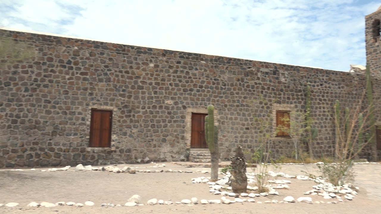 The rightward pan brings the edge of a stone bell tower into view beside the Mulege courtyard wall, with cacti, dry plants, and no visible people.