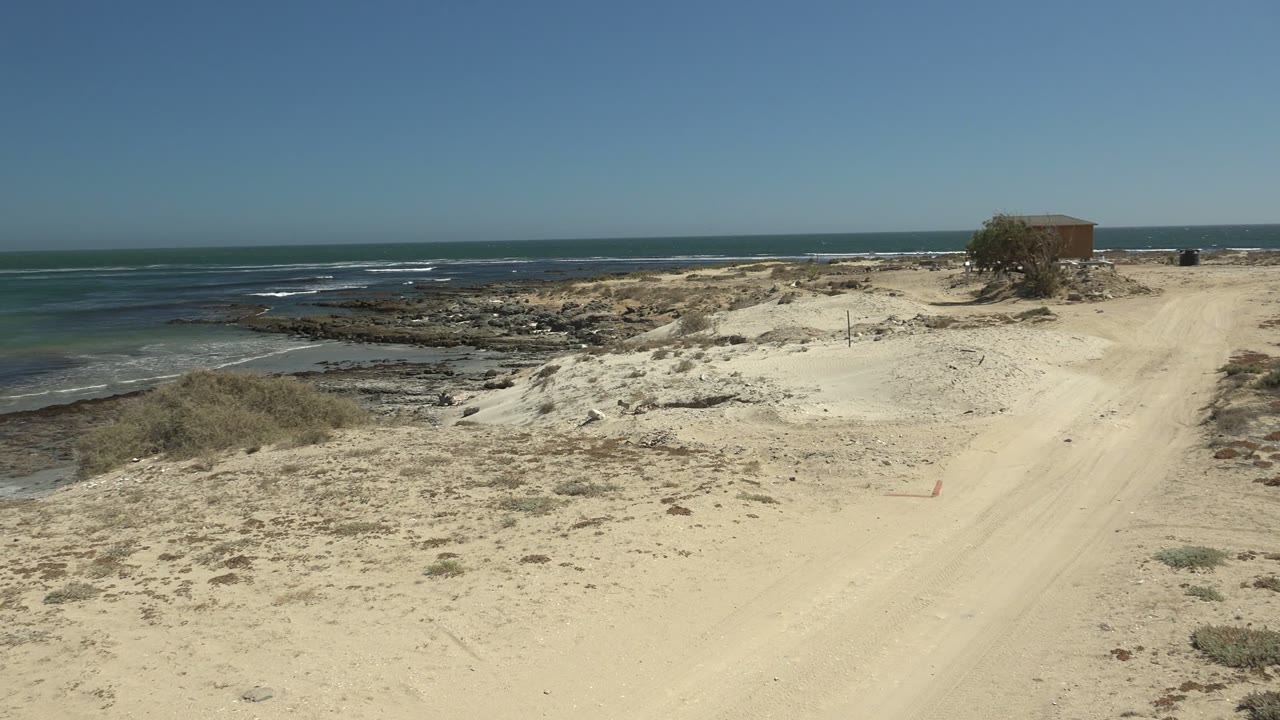La Bocana sandy beach path runs beside rocky ocean shore under a clear blue sky, with a small structure on the right and no visible people.