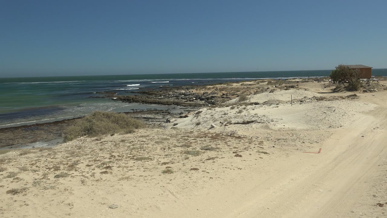La Bocana beach path crosses pale sand beside the ocean, with scattered scrub, rocky shore, a small structure near the upper right edge, and no visible people.