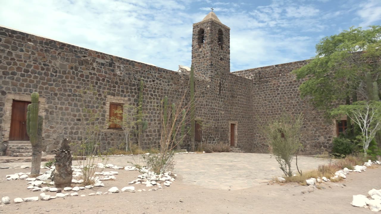Mulege, Baja California Sur stone building with a bell tower, cactus, white rock borders, sandy ground, trees, blue sky, and no visible people at the start of the clip.