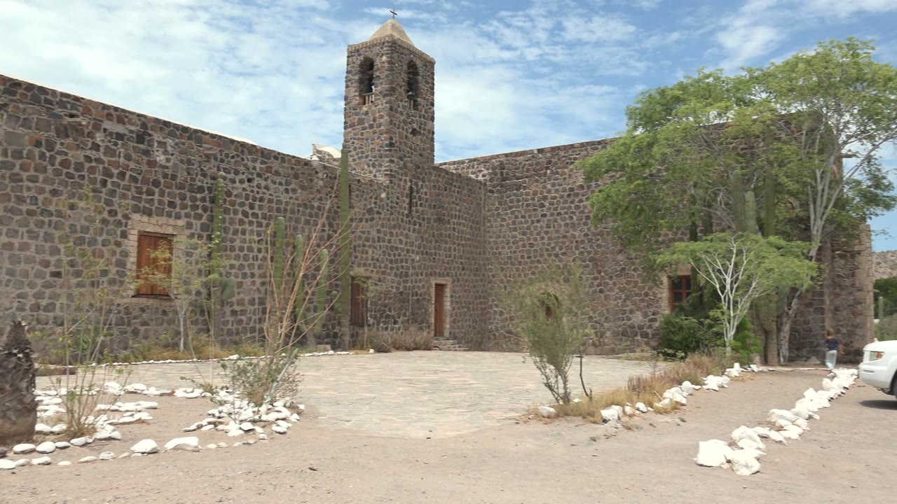 Mulege, Baja California Sur stone courtyard with bell tower, desert plants, white border rocks, trees, and the front edge of a white SUV beginning to enter at the right edge.