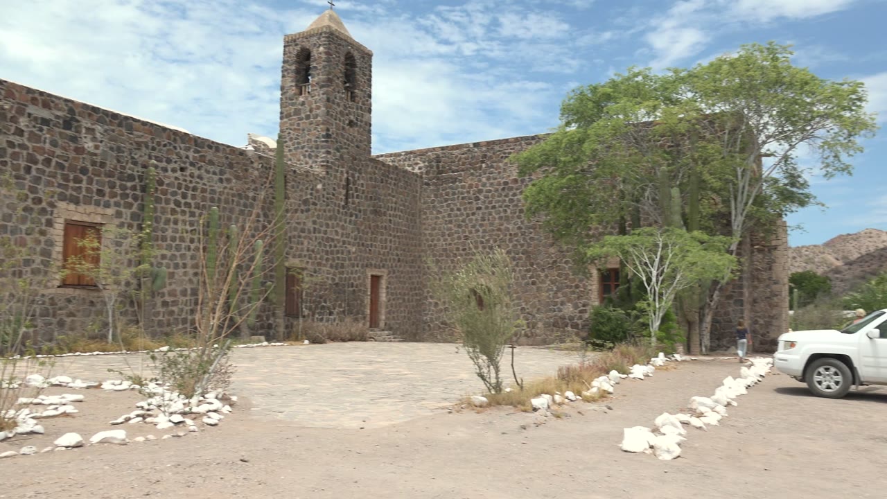 White SUV driving into a sandy stone courtyard beside trees and a bell tower.